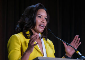 A woman in a bright yellow sports coat gestures with her hands while speaking at a lecturn.