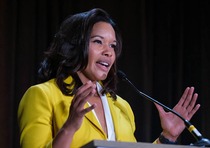 A woman in a bright yellow sports coat gestures with her hands while speaking at a lecturn.
